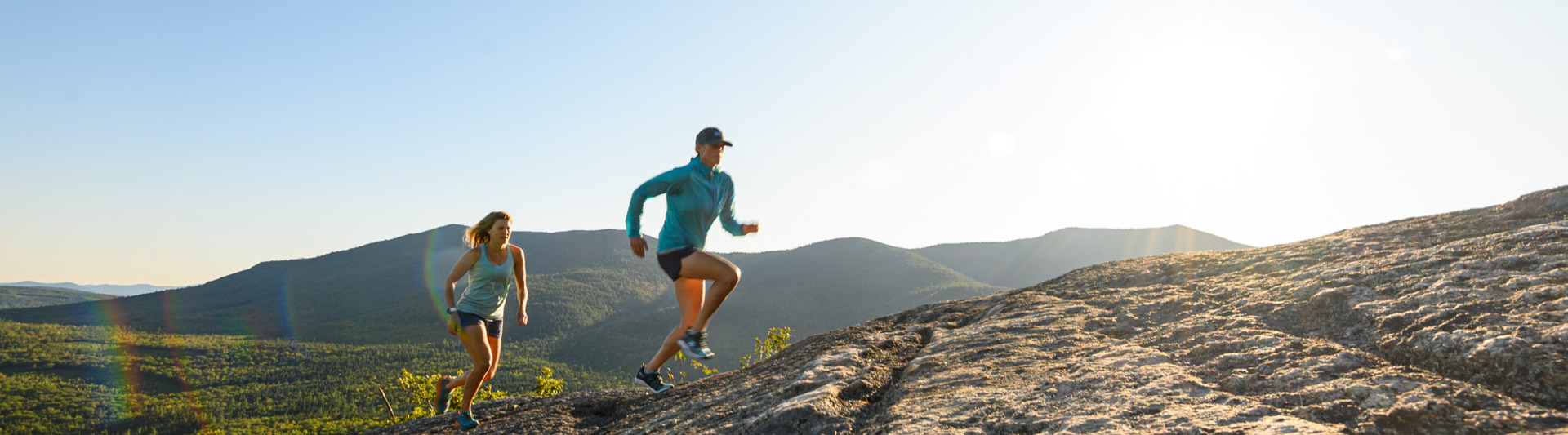 Two women trail running across a slab upa a mountain with the sun in the background